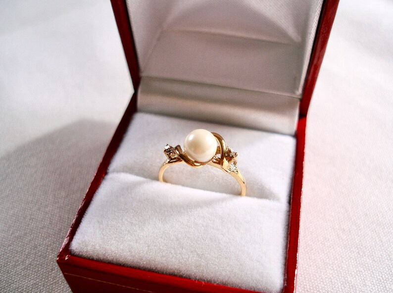 May include: A gold ring with a single pearl and two small diamonds set in a twisted band. The ring is displayed in a red velvet ring box.