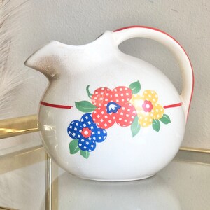 May include: White ceramic pitcher with a red rim and a floral design featuring red, yellow, and blue flowers with white polka dots.