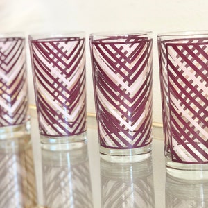 May include: Four clear glass tumblers with a geometric pattern of pink and burgundy lines. The glasses are arranged in a row on a gold surface.