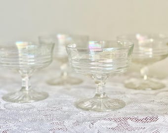 May include: Set of four clear champagne coupe glasses. Each glass features a round bowl with horizontal ridges and a stemmed base. The glasses are arranged on a white lace tablecloth, with a soft, diffused background.