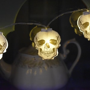 May include: A string of white fairy lights with paper skull shapes. Each skull has a small light inside, illuminating the skull from within. The lights are strung around a white teapot with a floral design.
