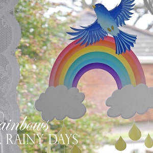 以下が含まれることがあります： 窓に青い鳥が乗ったカラフルな虹が飾られています。雲から黄色い雨粒が垂れ下がっています。画像の下には「Rainbows for Rainy Days」という文字が見えます。