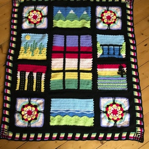 May include: A black crochet blanket with a colorful patchwork design. The blanket features various scenes, including mountains, cacti, and a window pane. The border is a striped pattern in pink, yellow, and green.
