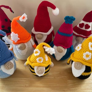 以下が含まれることがあります： さまざまな色の手作りの編み物ノームのコレクション。ノームは、赤、オレンジ、青、黄色の長いとがった帽子と白いアクセントが付いています。一部の帽子には、花柄やハートのデザインがあります。ノームは木製の表面に表示されます。