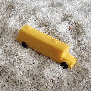 May include: A yellow toy school bus with black wheels, sitting on a bed of white sand.
