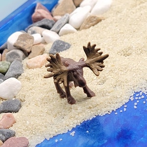 May include: A miniature brown moose figurine with large antlers standing on a bed of white sand with small brown and gray rocks.