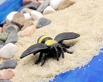 May include: A black and yellow bee figurine on a blue and white background. The bee is sitting on a bed of white sand with small white and brown rocks surrounding it.