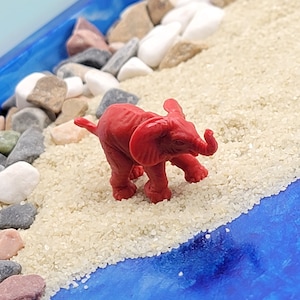 May include: A red plastic toy elephant figurine standing on a white sandy beach with small rocks and a blue background.