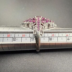 May include: A silver ring with a large, ornate design. The ring features a cluster of small, round, and rectangular pink gemstones. The ring is being measured on a metal ring sizer. The ring's band has an intricate, openwork design.
