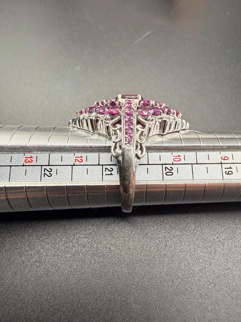 May include: A silver ring with a large, ornate design. The ring features a cluster of small, round, and rectangular pink gemstones. The ring is being measured on a metal ring sizer. The ring's band has an intricate, openwork design.