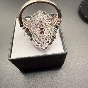 May include: A silver ring with a large, ornate setting. The setting is filled with small, pink gemstones and features a filigree design. The ring is displayed in a black box with a gray interior.