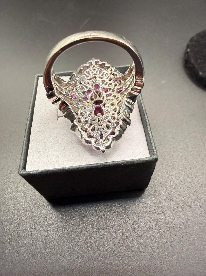 May include: A silver ring with a large, ornate setting. The setting is filled with small, pink gemstones and features a filigree design. The ring is displayed in a black box with a gray interior.