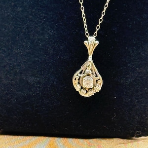 May include: A silver-toned metal pendant necklace featuring a teardrop-shaped frame with an openwork design. The pendant is set with a central round clear stone surrounded by smaller clear stones, suspended from a delicate silver-toned chain.
