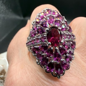 May include: A silver ring featuring a large, oval-shaped, deep red gemstone at the center, surrounded by numerous smaller, teardrop-shaped, and round, magenta-colored gemstones. The ring has an ornate, detailed setting.