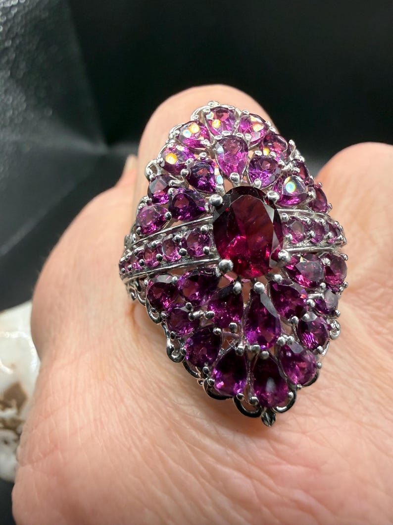 May include: A silver ring featuring a large, oval-shaped, deep red gemstone at the center, surrounded by numerous smaller, teardrop-shaped, and round, magenta-colored gemstones. The ring has an ornate, detailed setting.