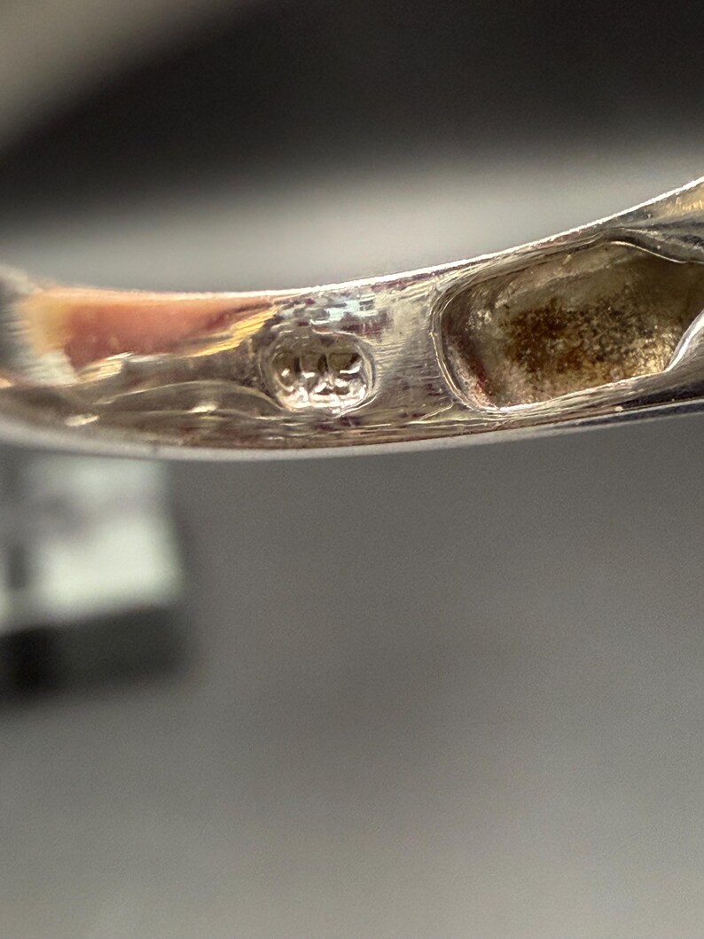 May include: Close-up of a silver ring with a hallmark stamp. The stamp reads "925". The ring's band is smooth and polished, reflecting the light. The ring is likely made of sterling silver.