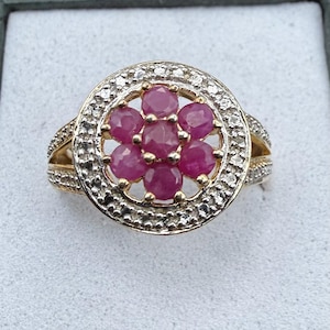 May include: A gold ring featuring a central cluster of seven round, faceted pink rubies arranged in a flower shape. The cluster is surrounded by a circular halo of small, clear pavé-set diamonds, with additional diamonds set along the split ring band.