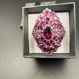 May include: A silver-toned ring with a large oval, deep pink gemstone at the center, surrounded by smaller, vibrant pink gemstones. The ring is set in a decorative, ornate design and is presented in a gray ring box.