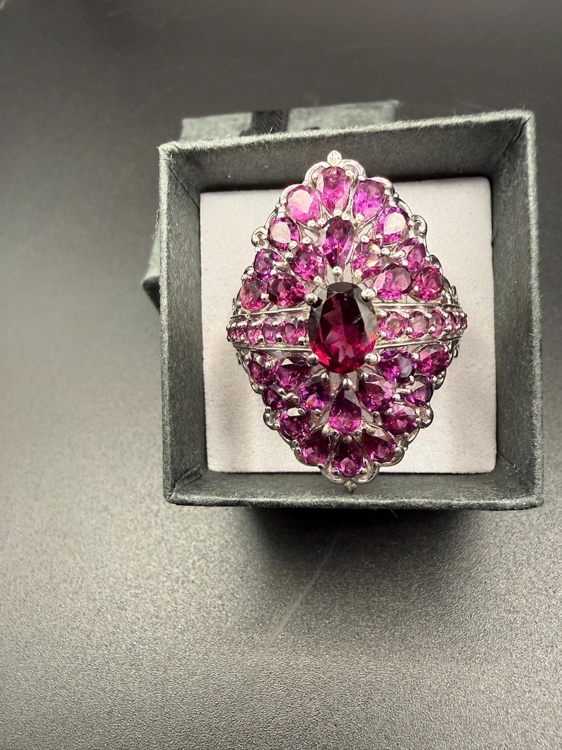 May include: A silver-toned ring with a large oval, deep pink gemstone at the center, surrounded by smaller, vibrant pink gemstones. The ring is set in a decorative, ornate design and is presented in a gray ring box.