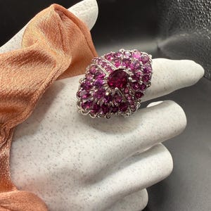 May include: A large, oval-shaped ring with a silver band and a cluster of small, round, and oval-cut pink gemstones. The center stone is a larger, deep pink gemstone. The ring is displayed on a white surface.