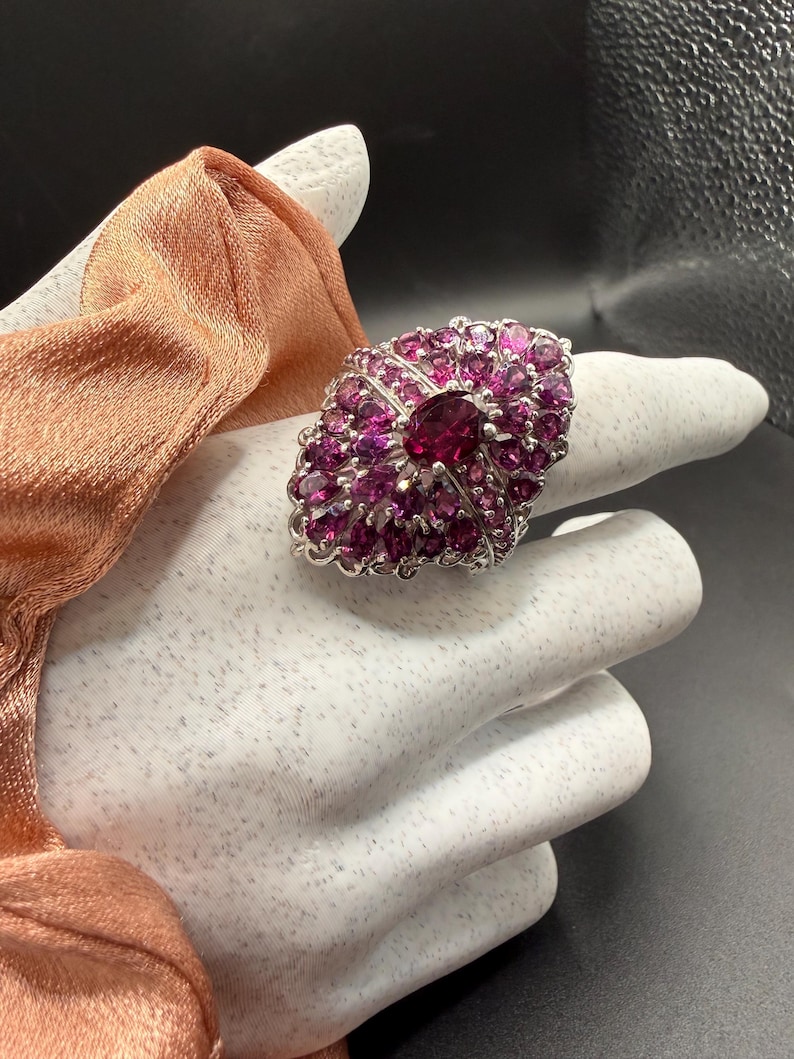 May include: A large, oval-shaped ring with a silver band and a cluster of small, round, and oval-cut pink gemstones. The center stone is a larger, deep pink gemstone. The ring is displayed on a white surface.