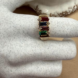 May include: A gold ring featuring four rectangular baguette-cut gemstones in red, blue, purple, and green, arranged vertically. Small, clear round accent stones are set between each colored gemstone on both sides of the band.