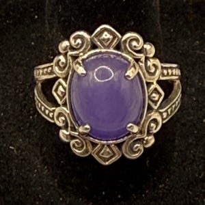 May include: A silver ring with an oval, lavender-colored stone. The ring has an ornate design with scrollwork and diamond-shaped accents. The ring is set against a black background.