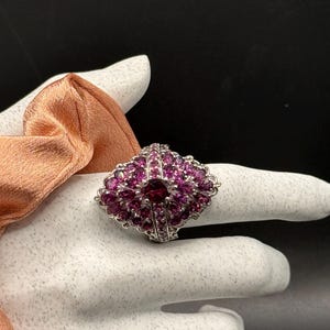 May include: A silver ring with a fan-shaped design, adorned with numerous small, round, pink gemstones and a larger, darker pink gemstone at the center. The ring is set against a white background.