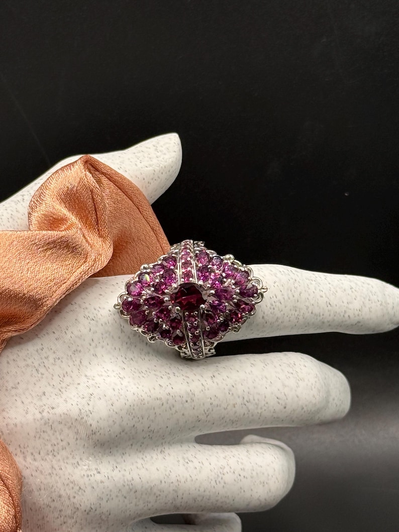 May include: A silver ring with a fan-shaped design, adorned with numerous small, round, pink gemstones and a larger, darker pink gemstone at the center. The ring is set against a white background.