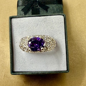 May include: A silver-toned ring featuring a large, oval-cut deep purple gemstone at the center, surrounded by a cluster of small, clear, sparkling stones set into the metal band, presented inside a dark green square box with a black ribbon.