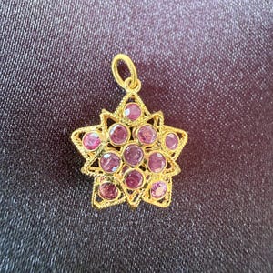 May include: A gold pendant shaped like a star with a textured, filigree-style frame. The center and points of the star are set with multiple round, faceted, deep pinkish-red gemstones arranged in a cluster, featuring a small gold bail at the top.