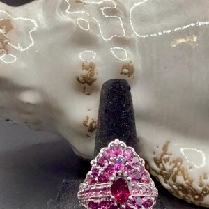 May include: A silver ring with a cluster of pink gemstones. The ring features a large oval-shaped center stone surrounded by smaller, petal-shaped stones. The ring is displayed on a black ring holder.