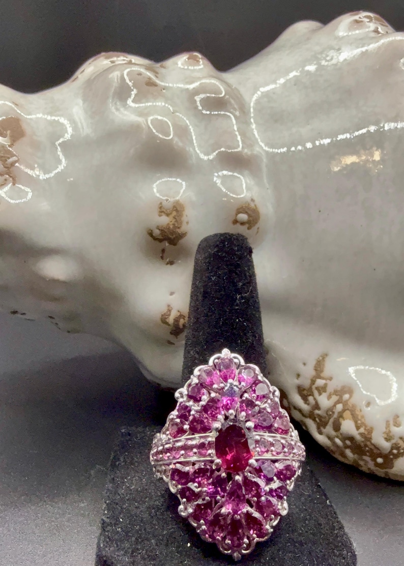 May include: A silver ring with a cluster of pink gemstones. The ring features a large oval-shaped center stone surrounded by smaller, petal-shaped stones. The ring is displayed on a black ring holder.