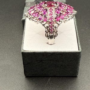 May include: A silver ring with a large, domed design. The ring is covered in numerous pink gemstones, with a larger, darker pink stone at the center. The ring is displayed in a gray gift box with a black ribbon.