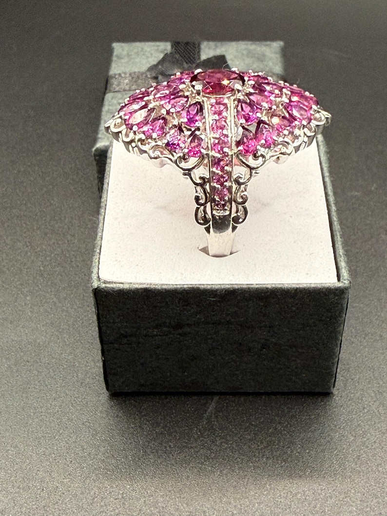 May include: A silver ring with a large, domed design. The ring is covered in numerous pink gemstones, with a larger, darker pink stone at the center. The ring is displayed in a gray gift box with a black ribbon.
