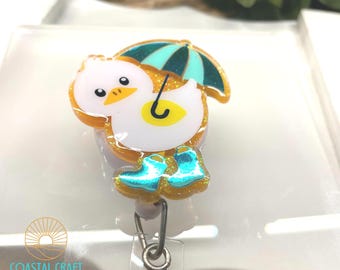Puede incluir: Un pato amarillo con un paraguas azul y botas de lluvia azules. El pato sostiene un paraguas amarillo y lleva botas de lluvia azules. El pato está hecho de resina y tiene detalles de purpurina. El pato está unido a un carrete de insignia retráctil.