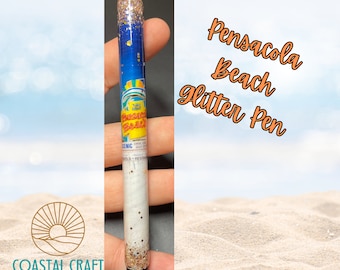 Puede incluir: Un bolígrafo de purpurina con un diseño azul y blanco que presenta el texto "Pensacola Beach" y una escena de playa. El bolígrafo está lleno de purpurina dorada y tiene una punta negra.