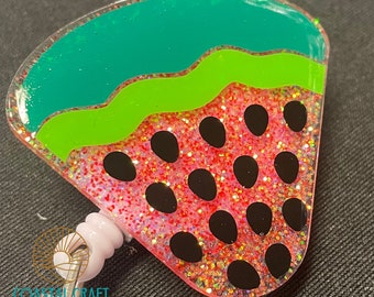 Puede incluir: Un portagafete en forma de sandía rosa y verde con semillas negras y purpurina. El portagafete tiene un respaldo de plástico transparente y un carrete retráctil.