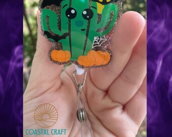 Puede incluir: Un portatarjetas con un cactus verde de ojos negros grandes, calabazas naranjas en la base, una luna amarilla y murciélagos negros. El diseño tiene un fondo de purpurina marrón con un detalle de telaraña blanca y una correa de plástico.