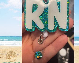 Puede incluir: Un portagafete de resina azul verdoso y blanco con las letras "RN" en blanco. El portagafete está unido a un carrete retráctil para gafetes.