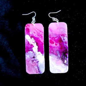 May include: A pair of long rectangular earrings with pink, white, and purple abstract patterns. The earrings have silver hooks.