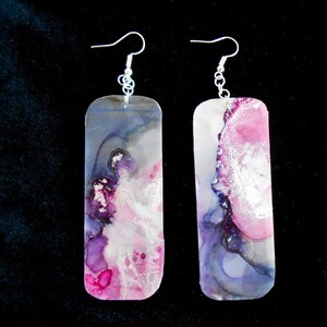 Peut inclure: Une paire de longues boucles d'oreilles rectangulaires avec un crochet argenté. Les boucles d'oreilles sont en résine et présentent un motif abstrait rose, violet et gris avec des accents argentés.