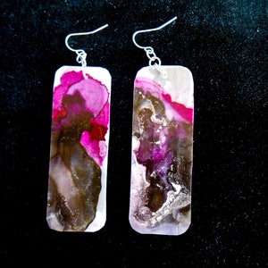 Könnte beinhalten: Ein Paar rechteckige Ohrringe mit einem abstrakten Design in Pink, Braun und Silber. Die Ohrringe sind aus Harz und haben silberne Haken.