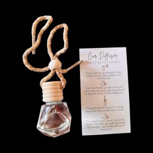 以下が含まれることがあります： 木製の蓋と装飾的なロープハンガーが付いた、透明で幾何学的な車の芳香剤。説明カードには「カーディフューザーの説明」と書かれています。芳香剤は吊り下げて、ボトルを逆さまにしてリフレッシュするように設計されています。