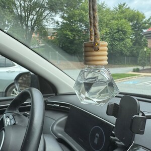 以下が含まれることがあります： 木製のキャップと編みロープが付いた、透明なファセットガラスの車用芳香剤。車の内装には、ハンドル、ダッシュボード、電話マウントが含まれています。背景には木々と曇り空が写っています。