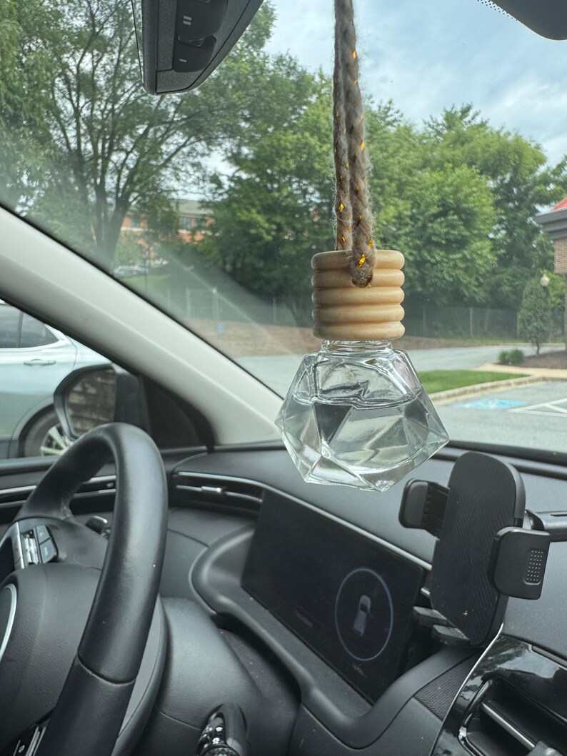 以下が含まれることがあります： 木製のキャップと編みロープが付いた、透明なファセットガラスの車用芳香剤。車の内装には、ハンドル、ダッシュボード、電話マウントが含まれています。背景には木々と曇り空が写っています。