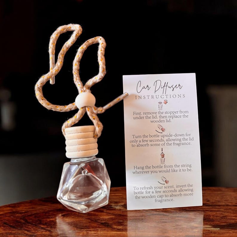以下が含まれることがあります： 木製の蓋と装飾的なロープハンガーが付いた、幾何学的な形状の透明な車用芳香剤ボトル。説明書が書かれたカードがボトルの隣にあります。カードのテキストは「カーディフューザーの説明」と書かれています。
