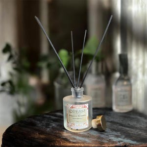 May include: A frosted glass aromatherapy diffuser with black reeds. The label reads "DREAM Bergamot Elements". A gold-colored cap sits next to the bottle. The diffuser is on a dark wooden surface.