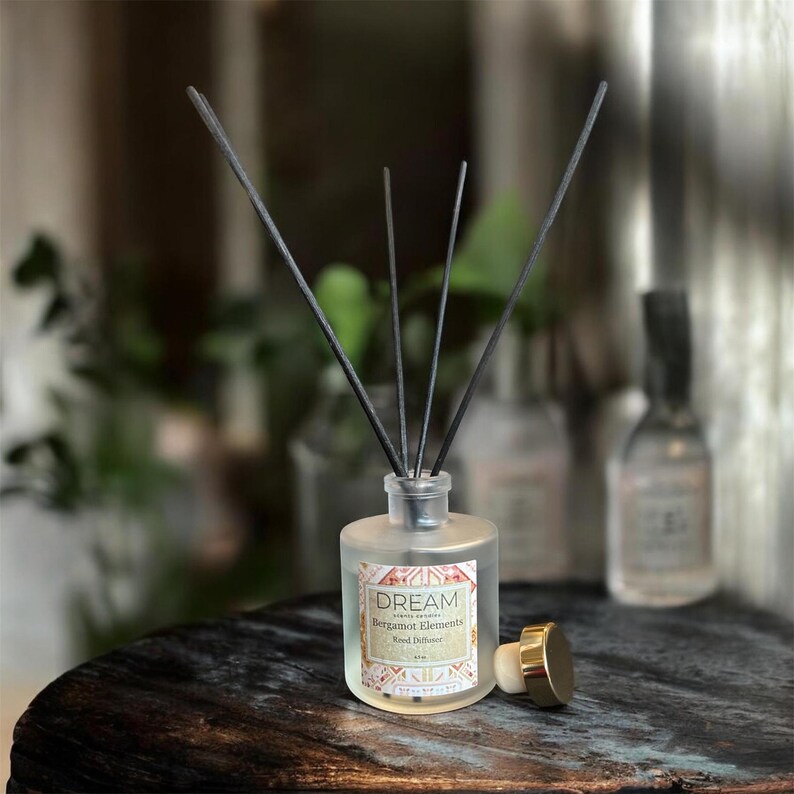 May include: A frosted glass aromatherapy diffuser with black reeds. The label reads "DREAM Bergamot Elements". A gold-colored cap sits next to the bottle. The diffuser is on a dark wooden surface.
