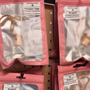以下が含まれることがあります： ピンク色のパッケージに入った車用芳香剤。木製のディフューザー、透明なクリスタル、金色の紐が特徴です。「Pineapple + Sage」と「Bergamot」のラベルが付いています。木製のディスプレイに吊り下げられています。